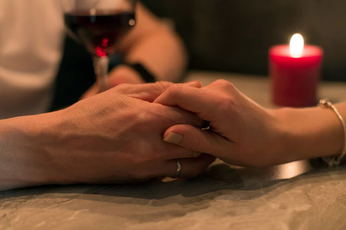 A romantic couple holding hands with a candle and wine glass in dim lighting, symbolizing love and intimacy.