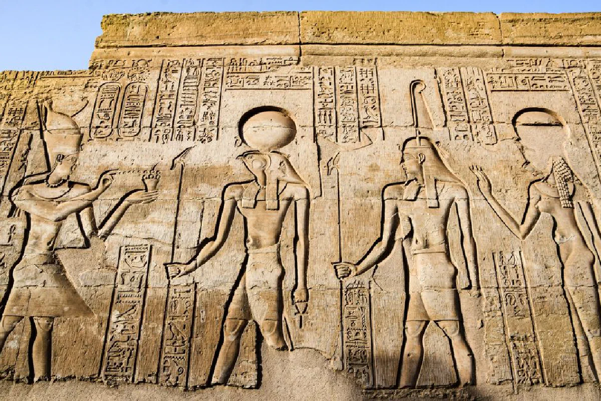 Detailed carvings of ancient Egyptian deities and hieroglyphs on a temple wall in bright daylight.