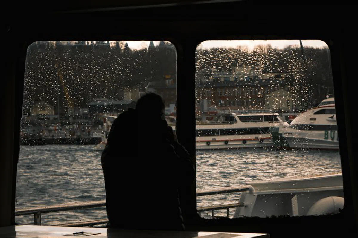 A silhouette captured inside a boat with a rainy window overlooking the city and docked boats.