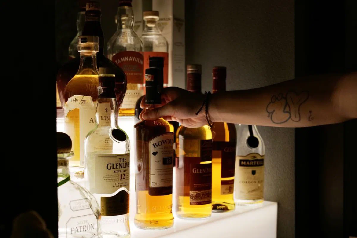 A tattooed hand selects a whiskey bottle from an illuminated shelf, showcasing various liquor options at a bar.