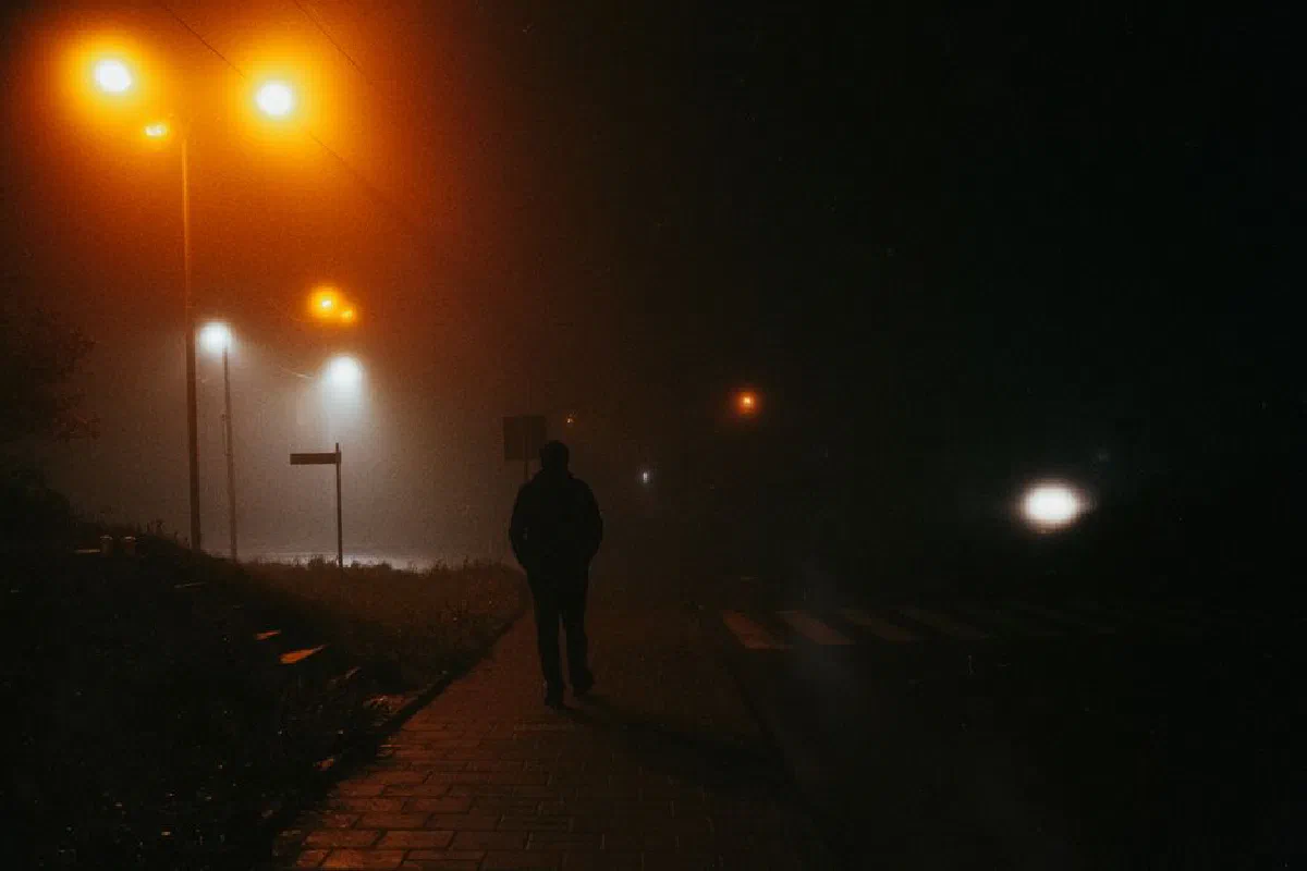 A lone silhouette walks on a foggy street at night, illuminated by streetlamps, creating a moody atmosphere.