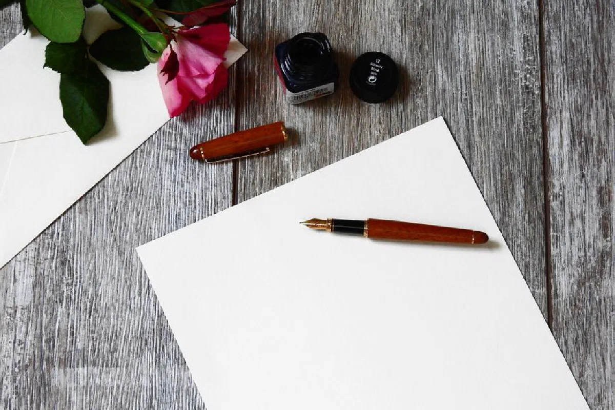 Flat lay image of a rose, fountain pen, and paper on a rustic wooden table.