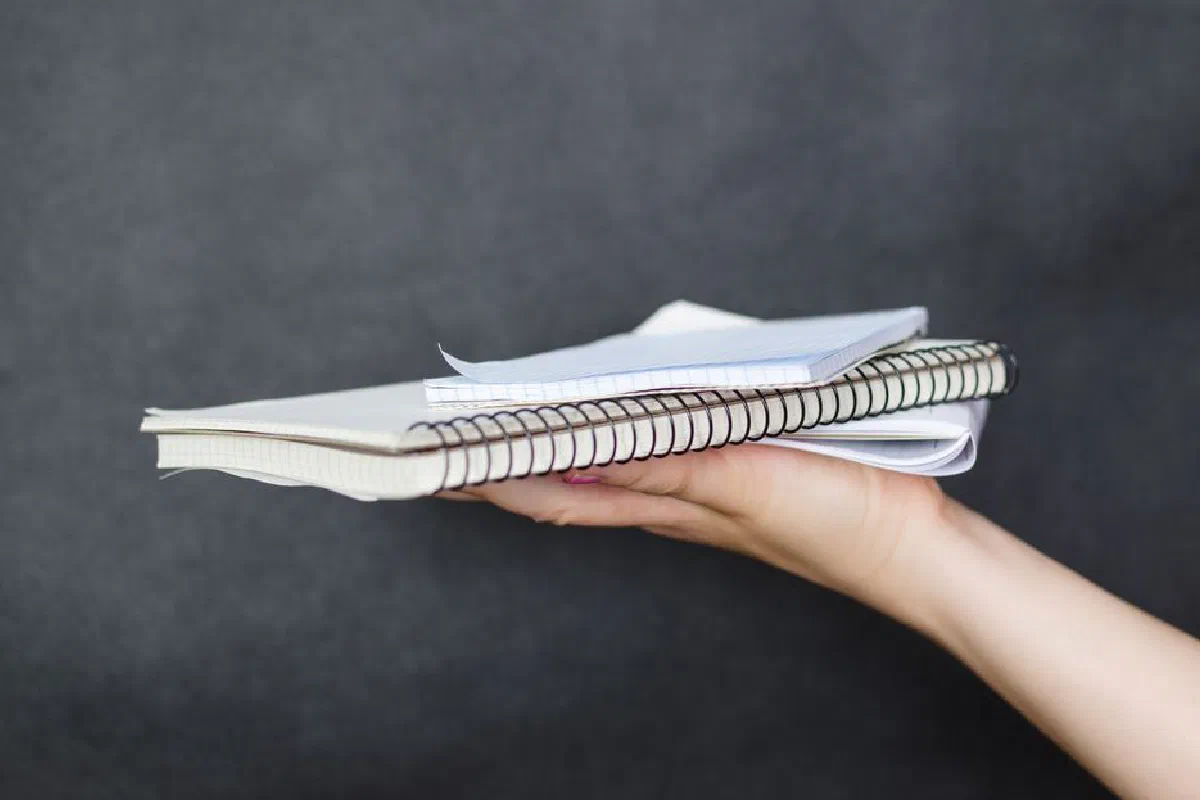 Close-up image of a woman's hand holding a stack of spiral-bound notebooks and papers against a dark background.