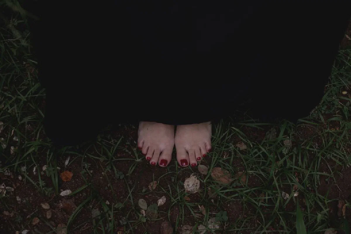 Close-up of bare feet wrapped in a dark cloak standing on grass, evoking mystery.