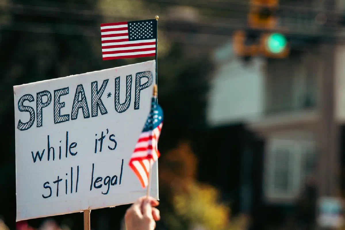 Protest sign with the message and American flag at a rally, highlighting free speech themes.