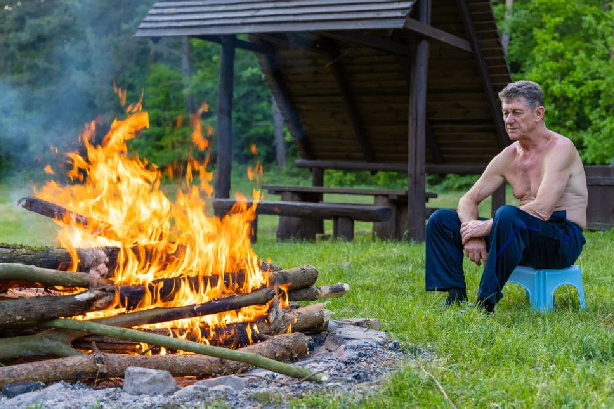 A shirtless man sitting by a campfire at a forest campsite in Poland.