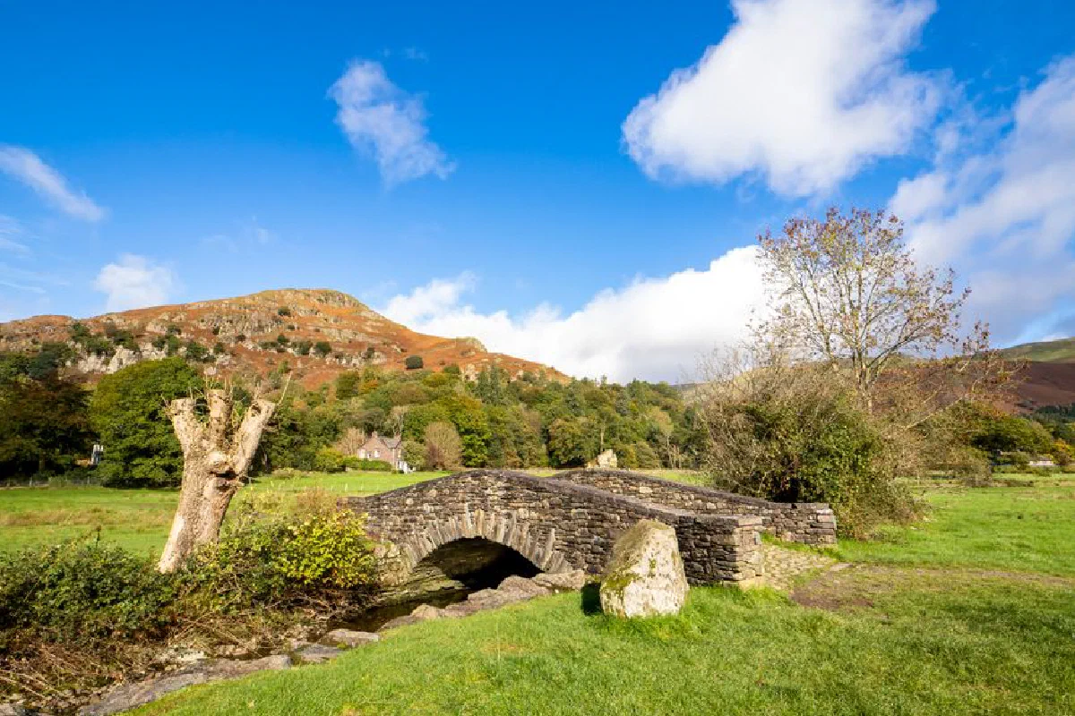 Charming stone bridge over a stream in a picturesque rural landscape with mountains and blue sky.