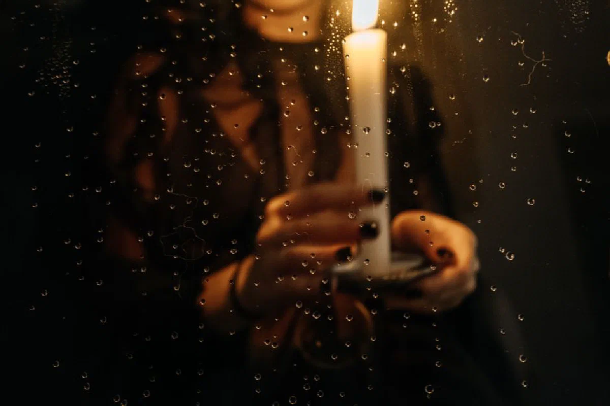 A mysterious, moody candlelit scene with raindrops on a glass window and a person holding a candle.