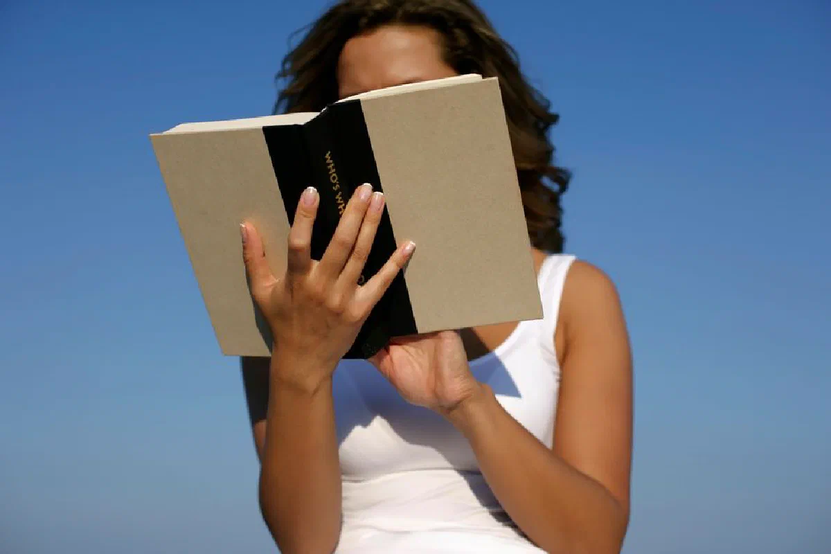 A woman in a white dress reads a book against a clear blue sky on a sunny day, enjoying leisure time outdoors.