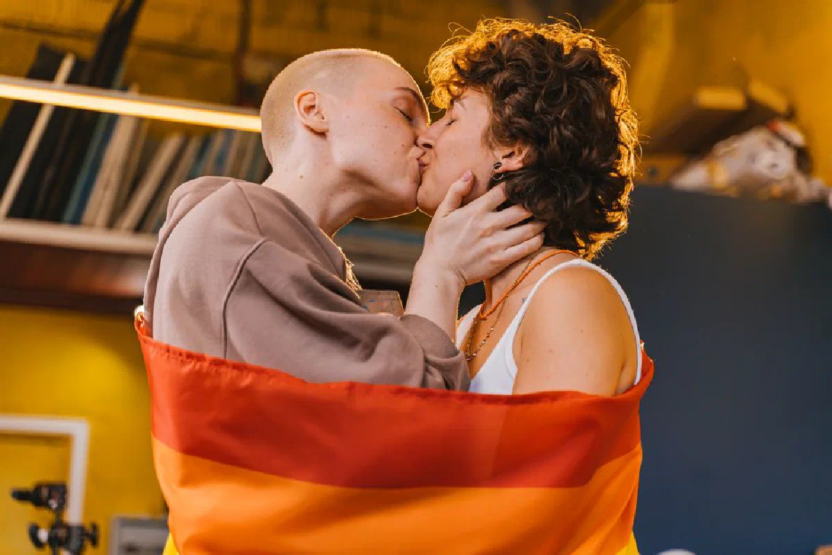 Two women embracing and kissing wrapped in a pride flag, expressing love and affection.