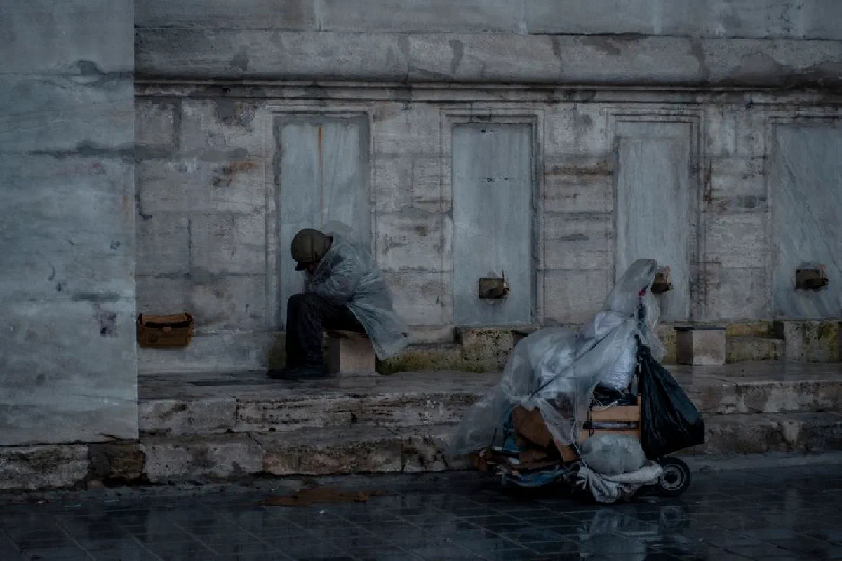 A homeless man in a rain poncho sits beside belongings on a rainy street, portraying urban hardship.