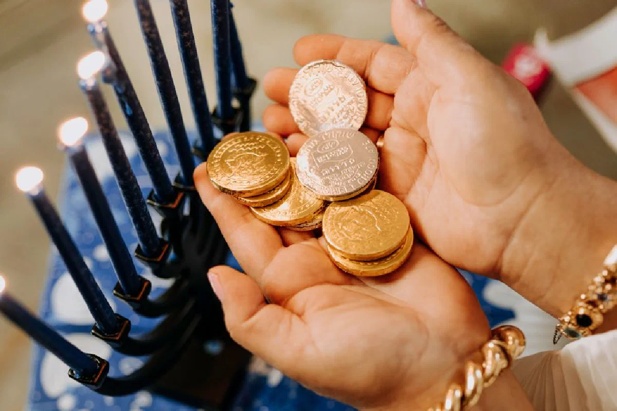 Hands holding gelt coins beside a lit menorah, celebrating Hanukkah indoors.