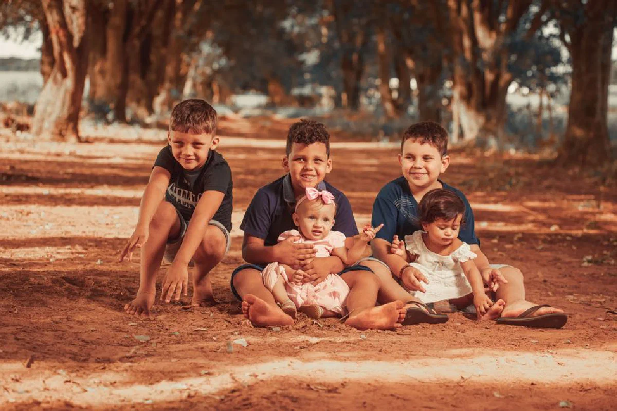 A group of cheerful children sitting barefoot on a dirt path surrounded by trees.