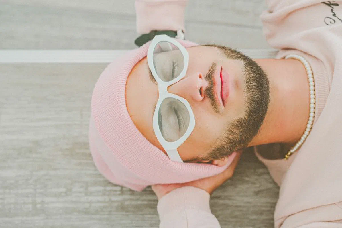 A bearded man relaxes outdoors, wearing a pink beanie and sunglasses.
