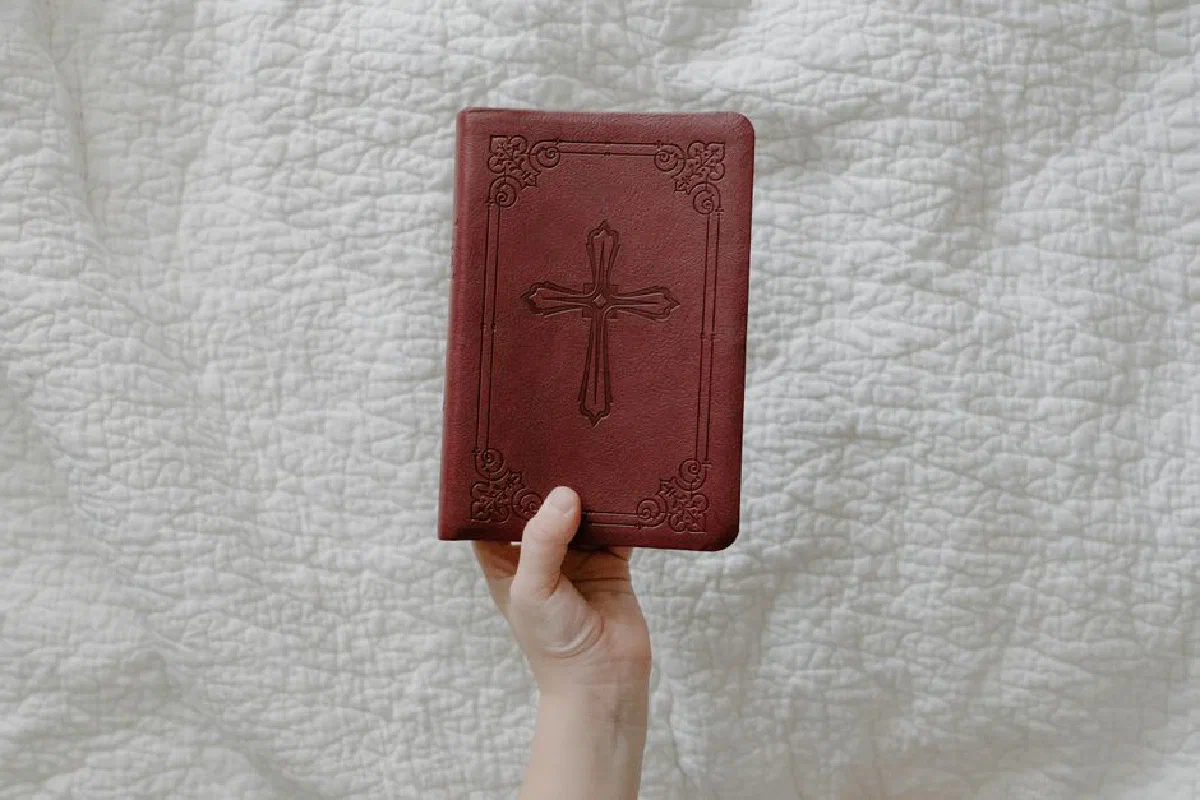 Close-up of a hand holding a leather-bound Bible with cross on a white quilt.