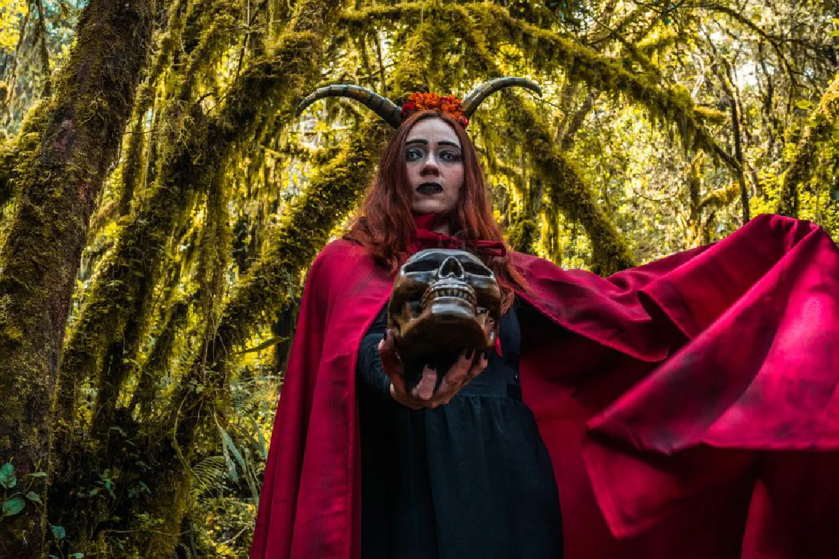 A mysterious woman in a red cape and horns holds a skull in a mystical forest.