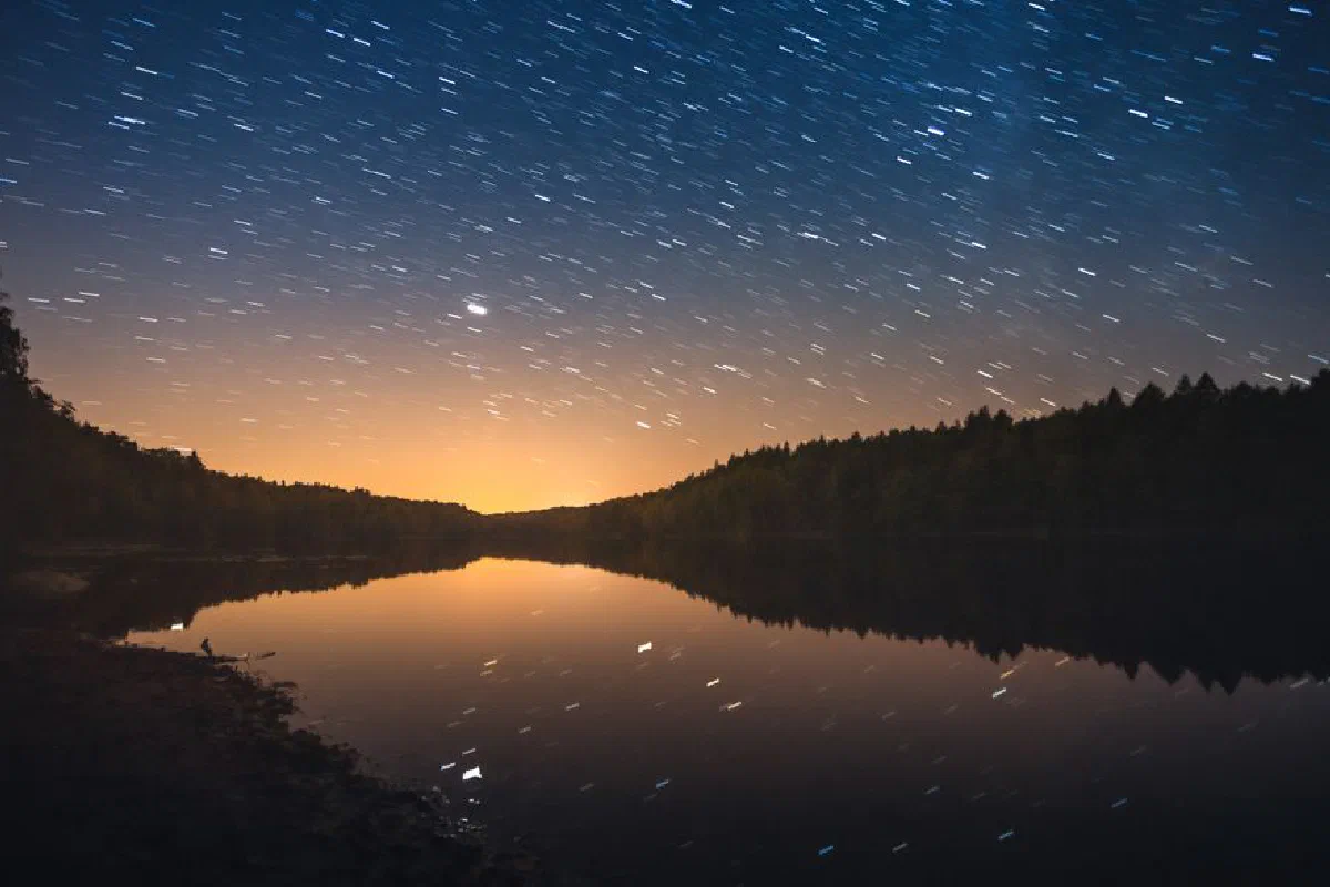 A mesmerizing night sky with star trails reflecting on a calm lake surrounded by trees.