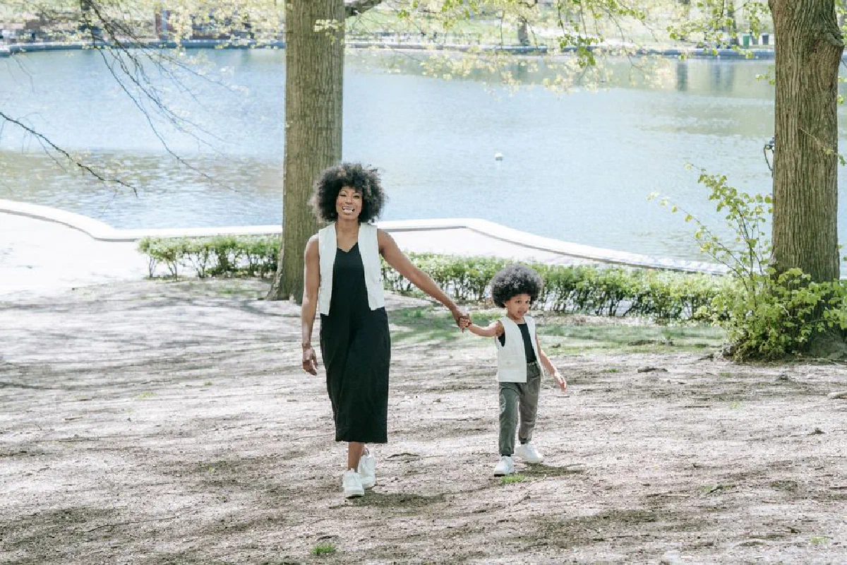 A mother and her child walk hand in hand by a peaceful lakeside in a sunny park.