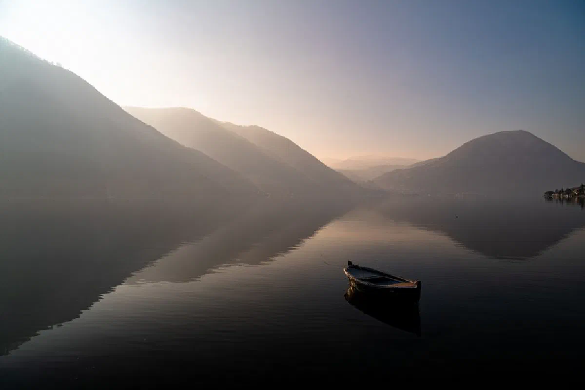 Calm water with a lone boat at sunrise, surrounded by misty mountains.