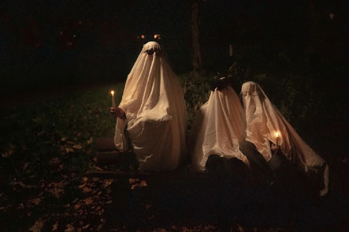 Three ghostly figures with candles on a bench in a dark, eerie park.