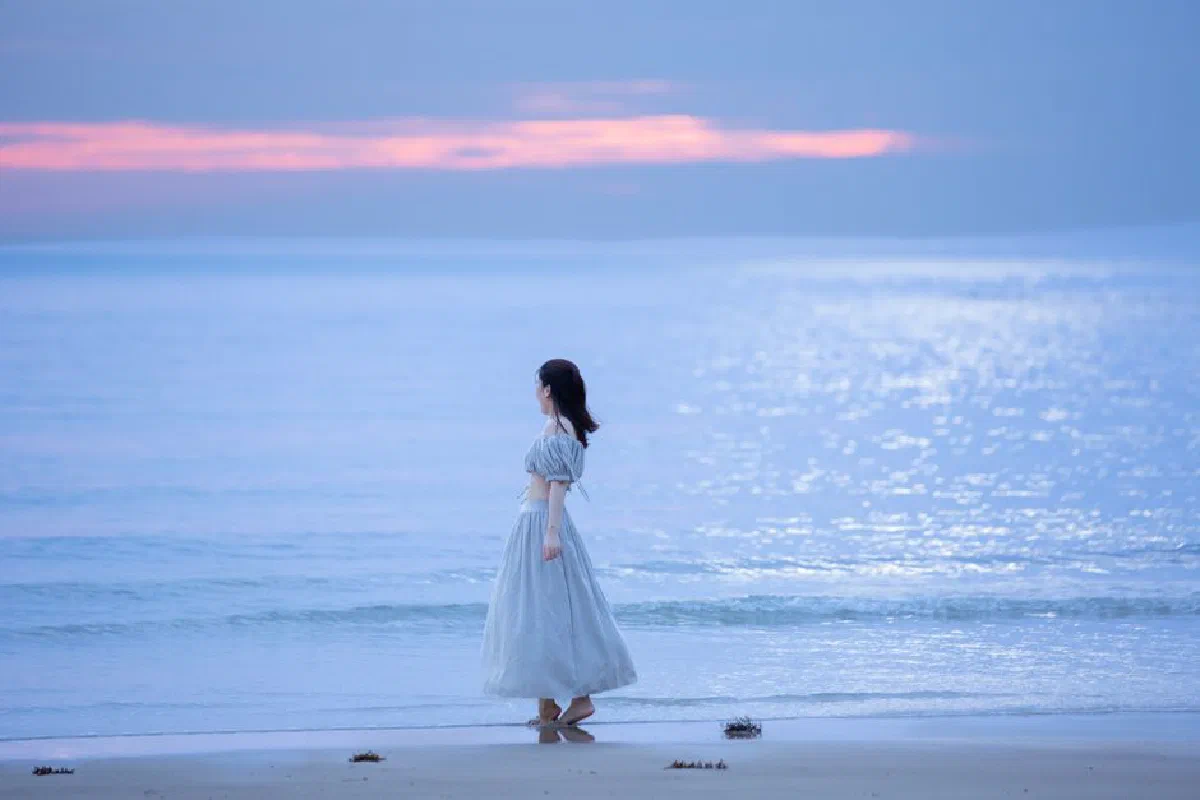 A woman in a flowing white dress walks along a serene beach during twilight, creating a calm, peaceful atmosphere.