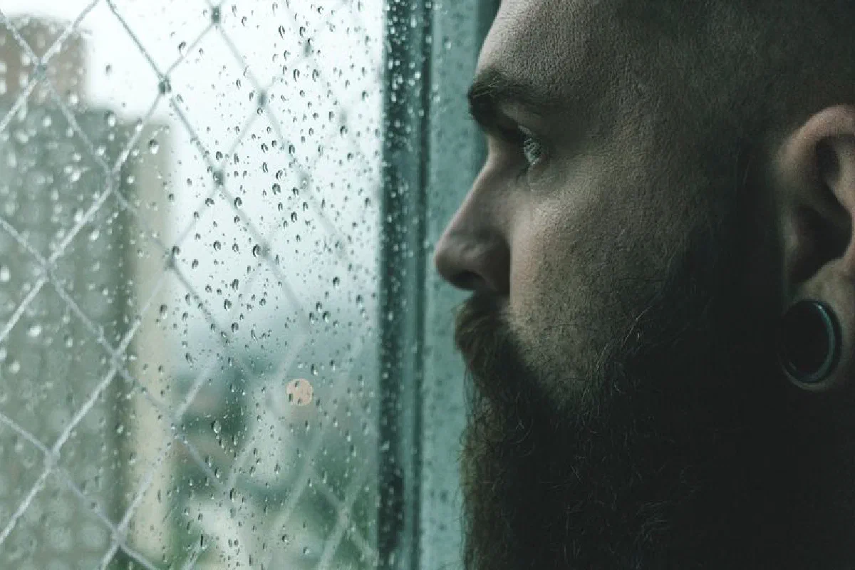 Close-up of a man with a beard looking thoughtfully through a rain-spattered window.