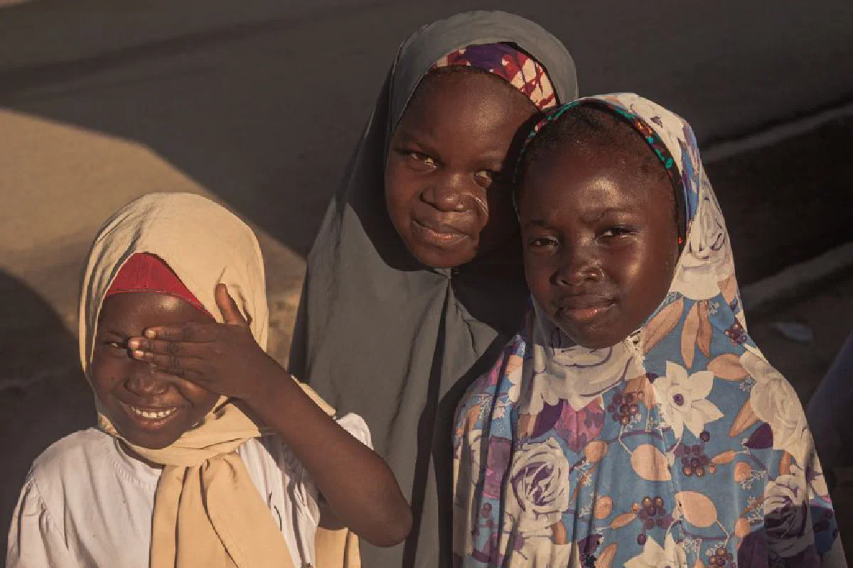 Three children wearing vibrant hijabs smile warmly in an outdoor setting.