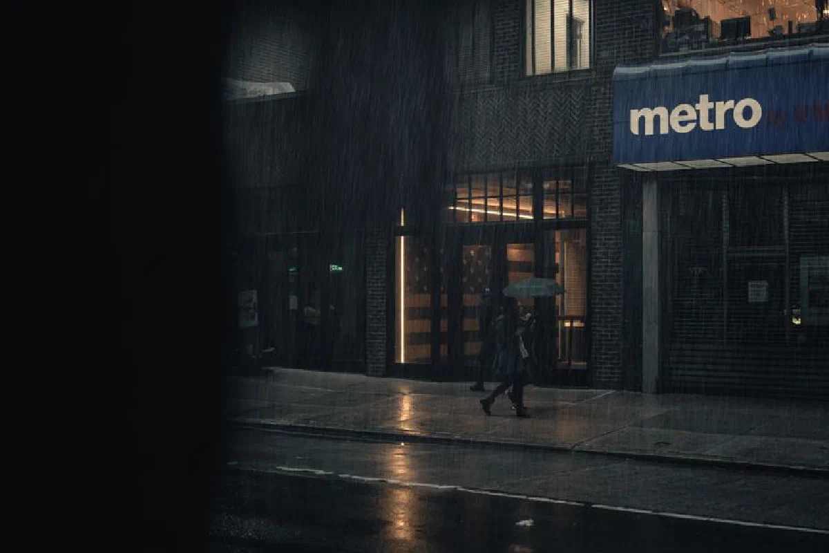 A person walks on a rainy city sidewalk at night, illuminated by streetlights, holding an umbrella.