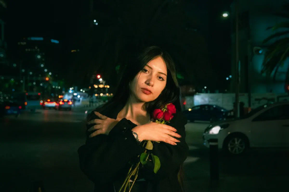 A thoughtful woman embraces red roses on a city street at night, illuminated by urban lights.