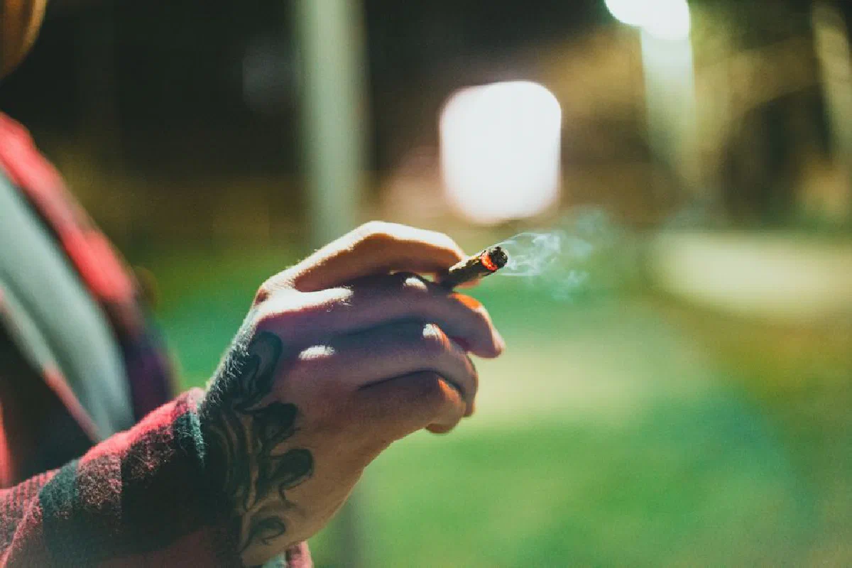 Close-up of a tattooed hand holding a lit joint outdoors at night with a blurred background.