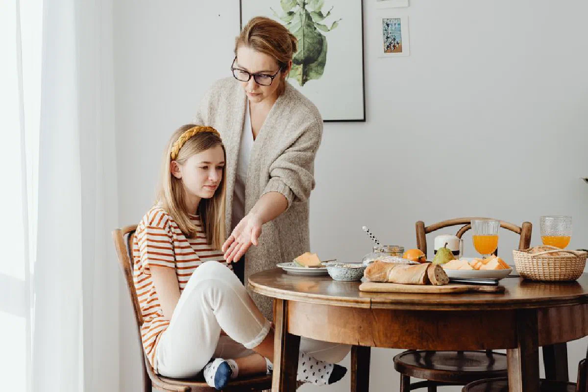 A mother guides her teenage daughter in dining etiquette at a cozy indoor setting.