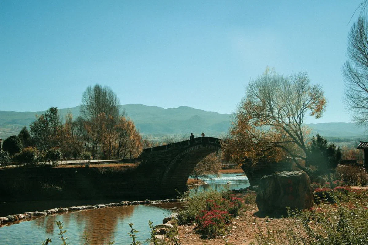 A tranquil summer scene featuring a stone bridge over a quiet river surrounded by lush trees.