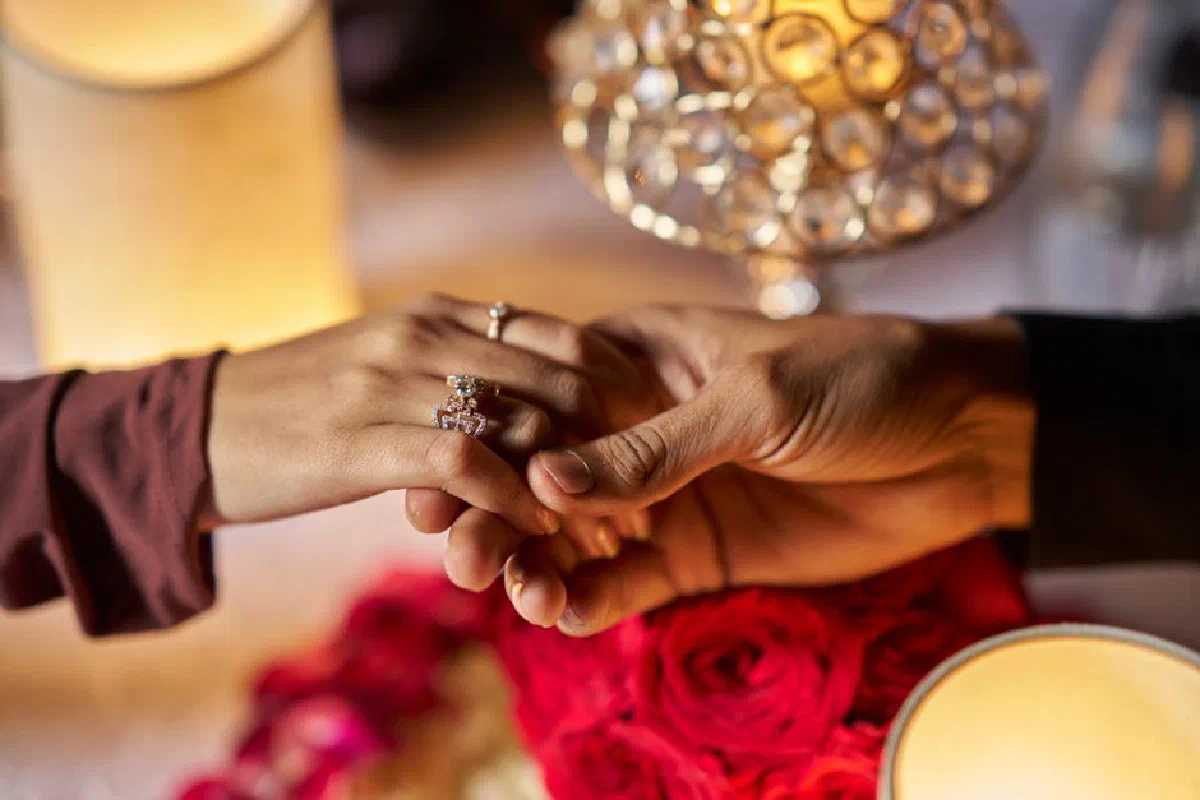 Close-up of a couple holding hands with engagement rings beside romantic candles and roses, symbolizing love and commitment.