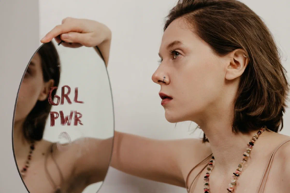 A young woman gazes into a mirror with 'GRL PWR' written on it, emphasizing empowerment.