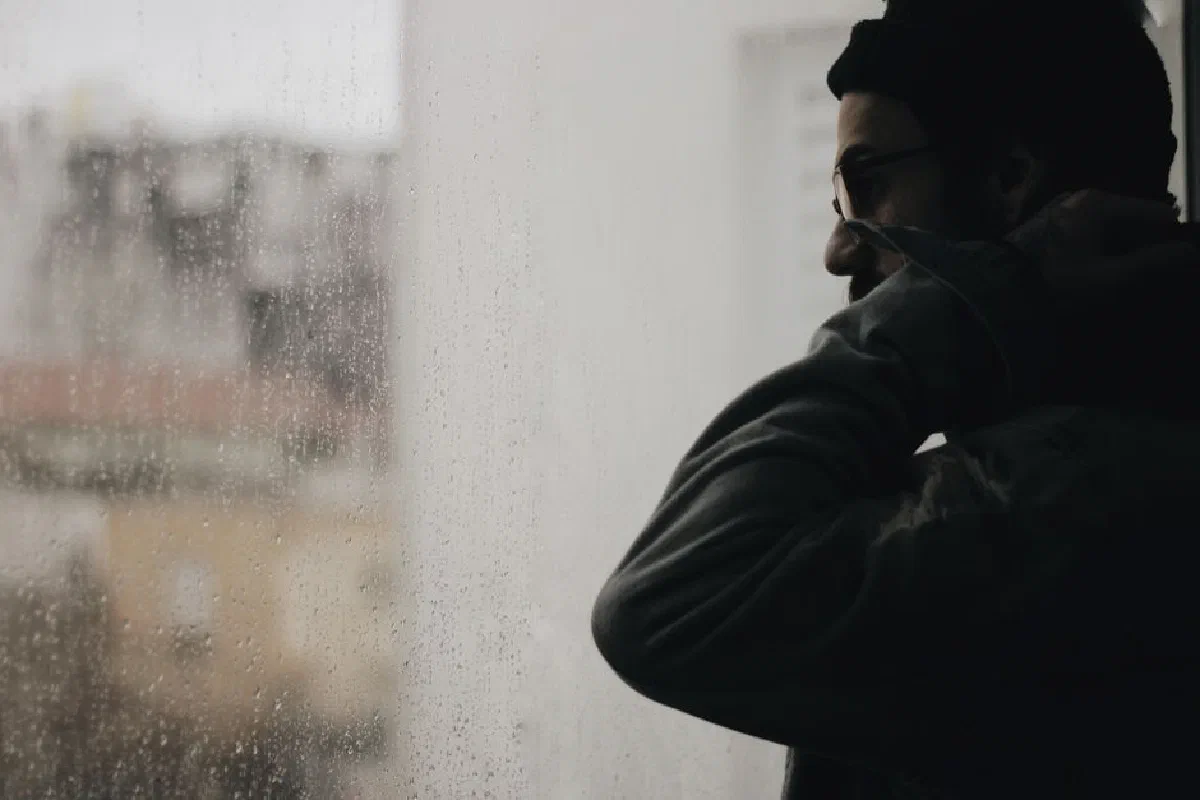 A man in a beanie gazes outside through a rainy window, creating a moody atmosphere.