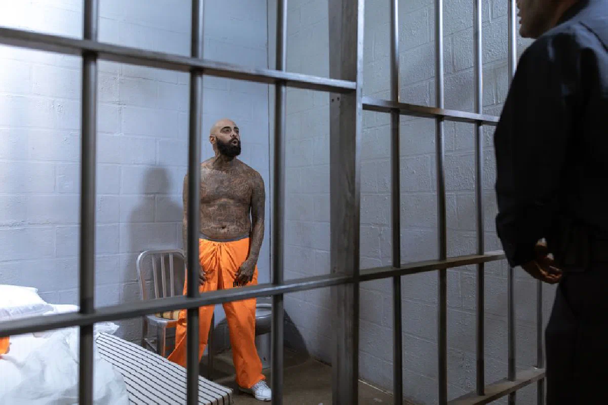 A tattooed prisoner stands behind bars in an orange jumpsuit under a guard's watch.