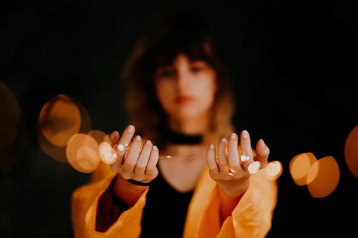 A young woman in a yellow raincoat holds string lights, creating a soft bokeh effect.