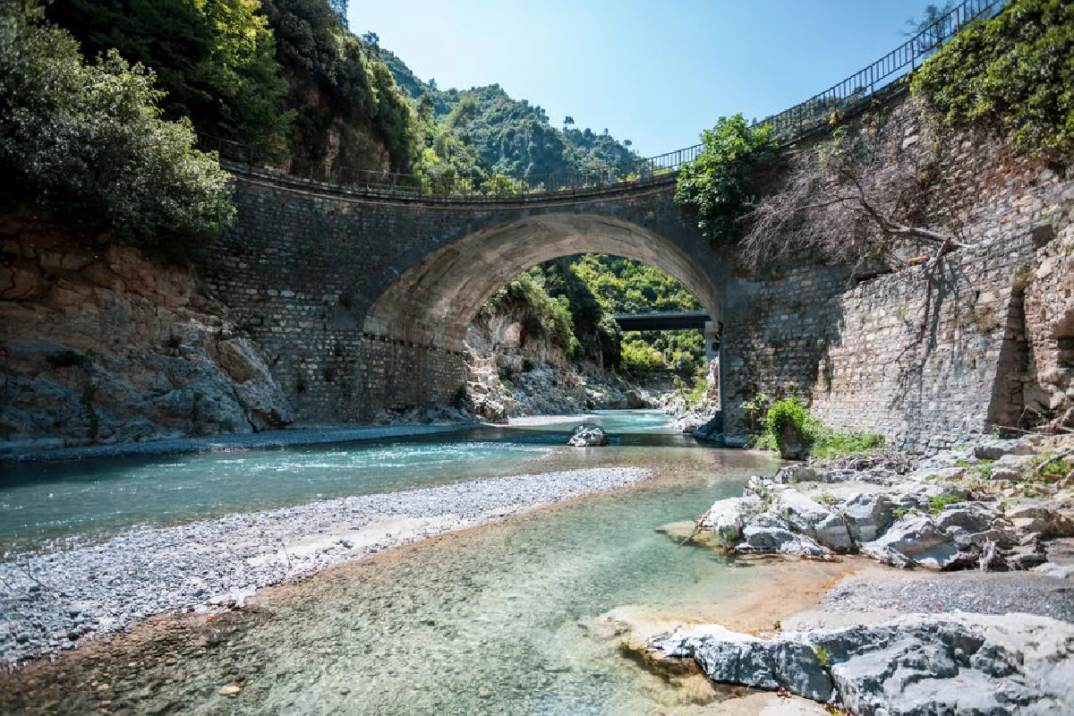 A scenic view of an old stone bridge over a clear river in a mountainous landscape.