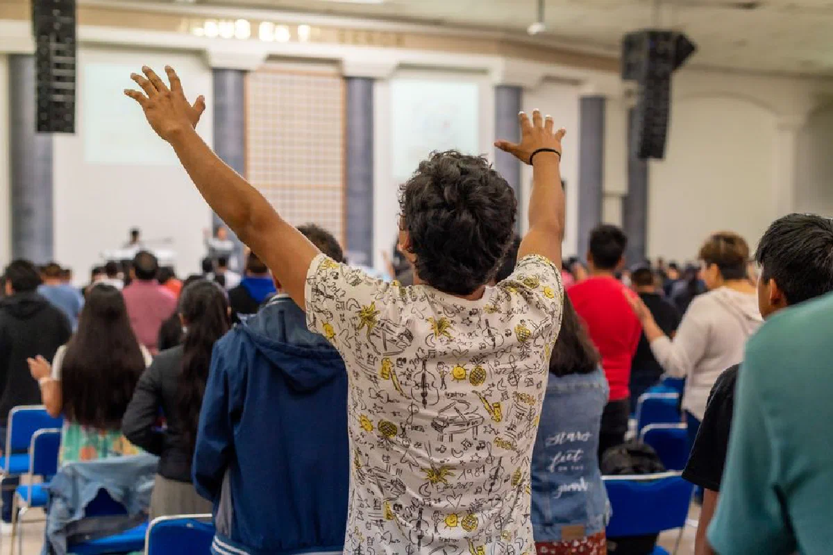 A diverse group of people raises hands in worship inside a large hall during a service.