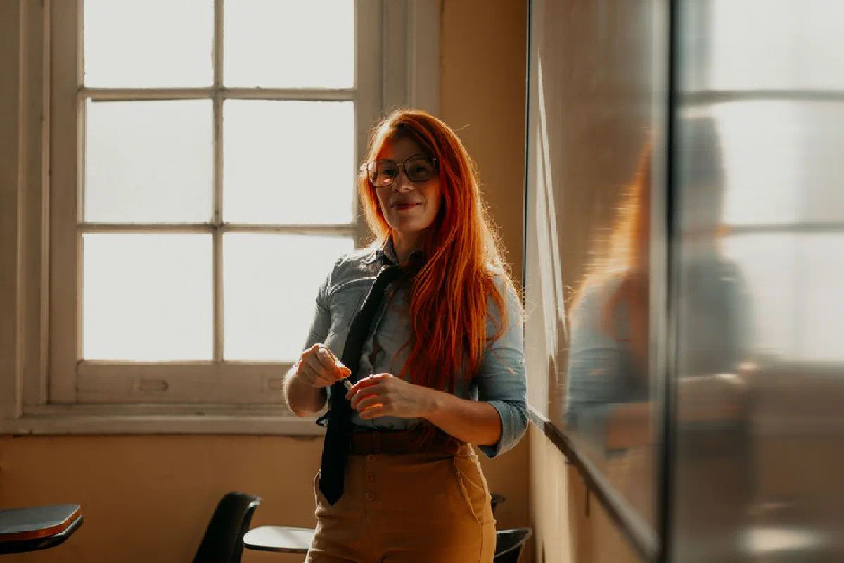 Smiling female teacher in Buenos Aires classroom during daylight, standing by the window with a chalkboard.