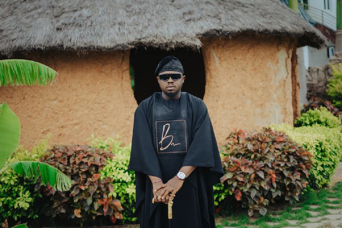 African man in traditional clothing poses by a rustic hut outdoors.
