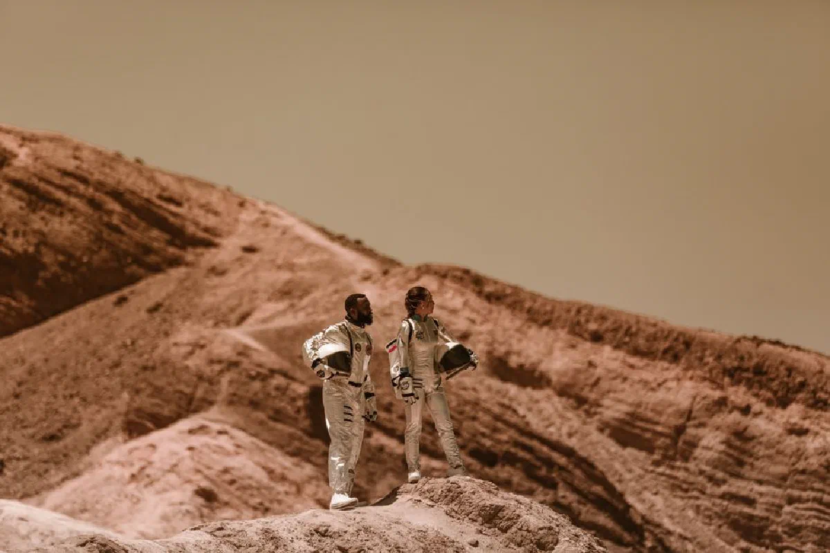 Astronauts exploring a desert-like terrain with rugged hills, wearing space suits.