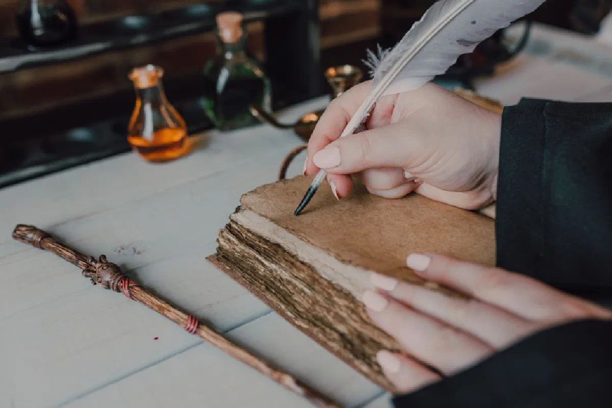 Close-up of hands writing with a quill in a spellbook, featuring magical items and a wand.