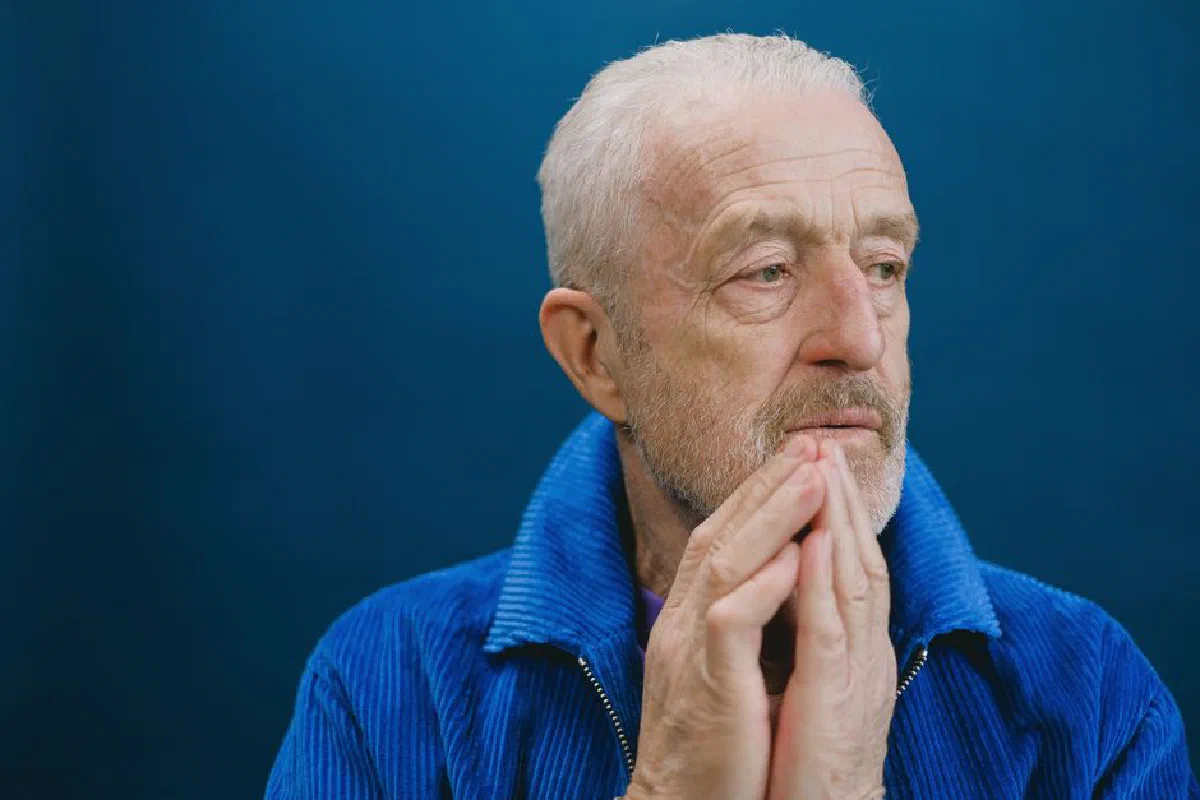 Elderly man with white hair and beard in a blue jacket, hands together, against a blue background.