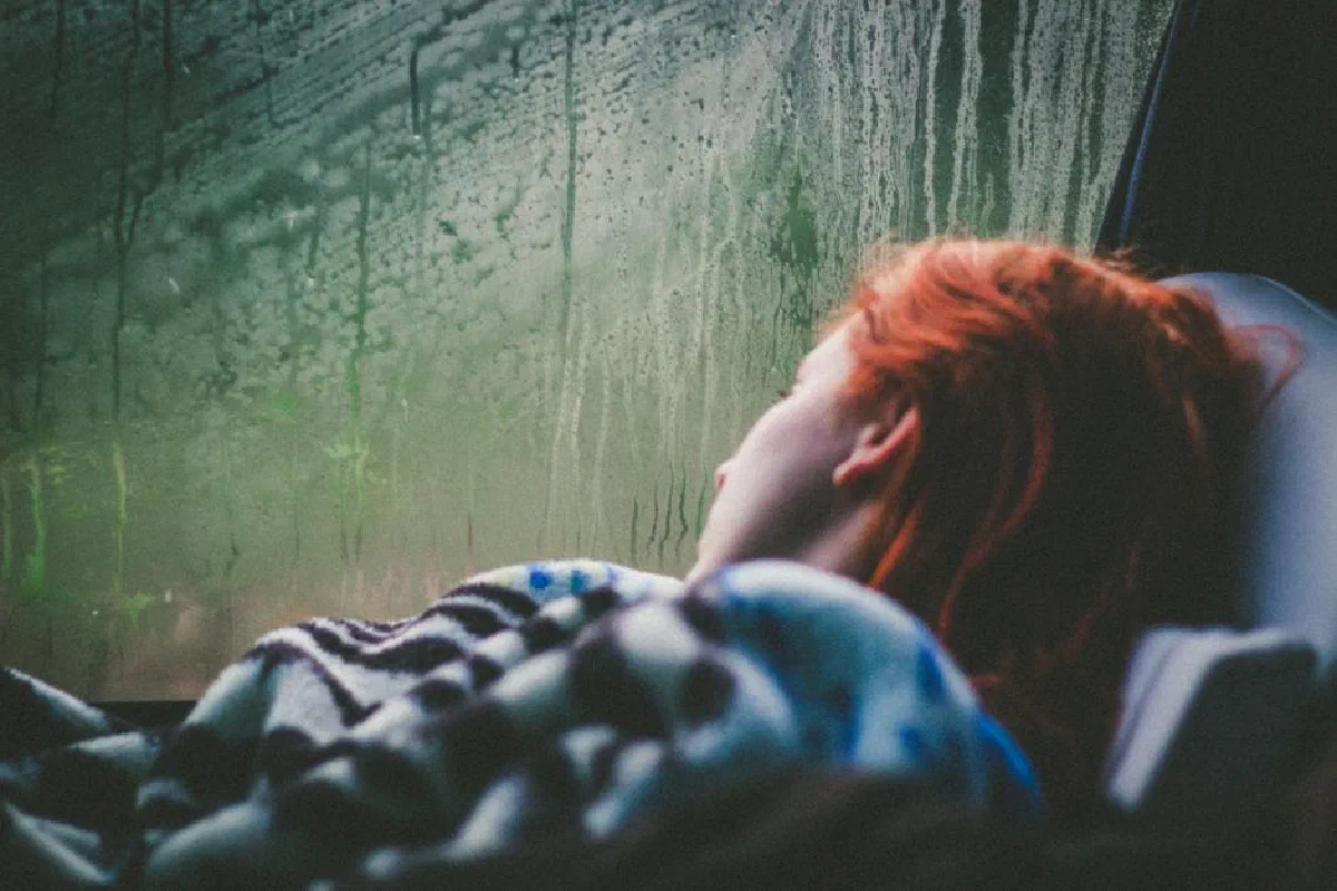 Woman with red hair relaxing, lying on a blanket by a wet window, exuding calm.