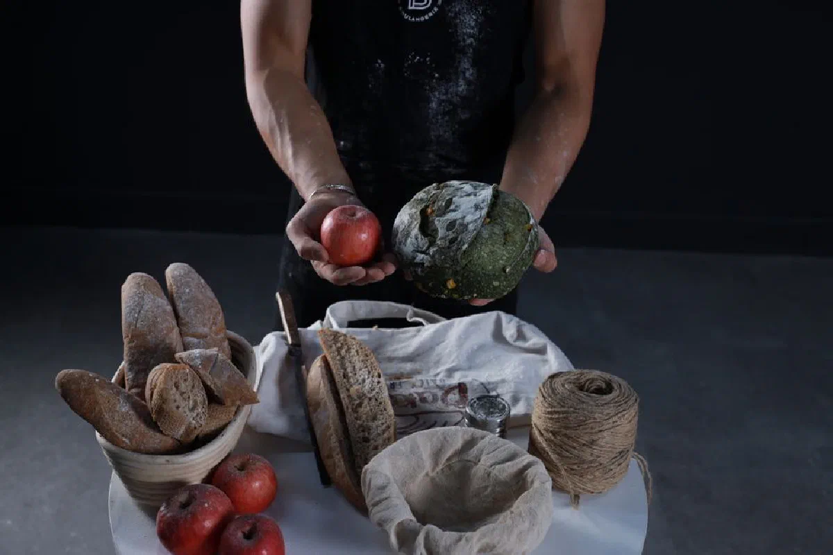 A man displays artisanal bread and a red apple with rustic food items on a table.