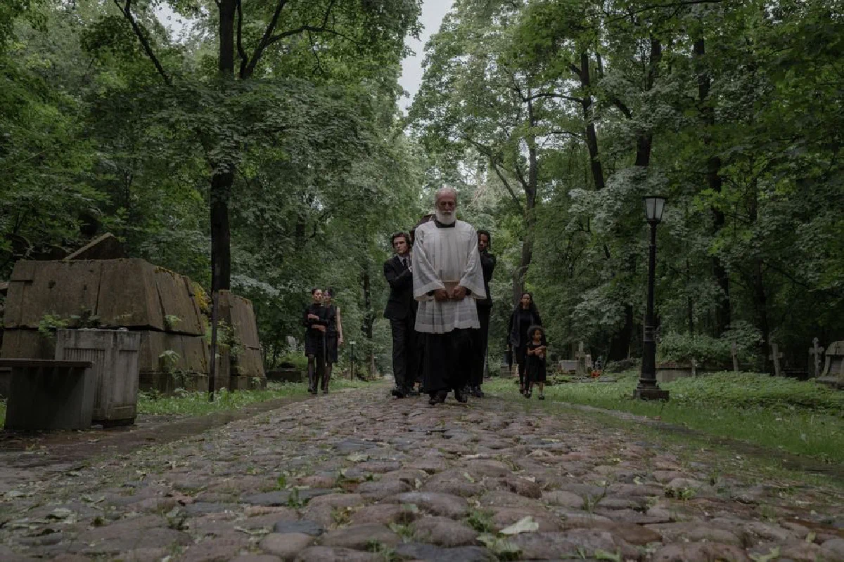 A group of mourners dressed in black walk through a peaceful cemetery path surrounded by lush trees.