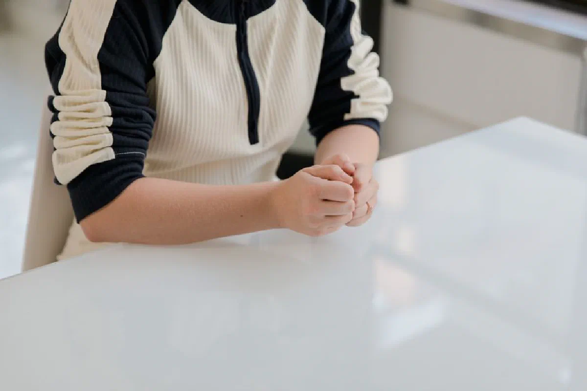 Close-up of a woman seated indoors, folding her hands on a table, suggesting contemplation or waiting.