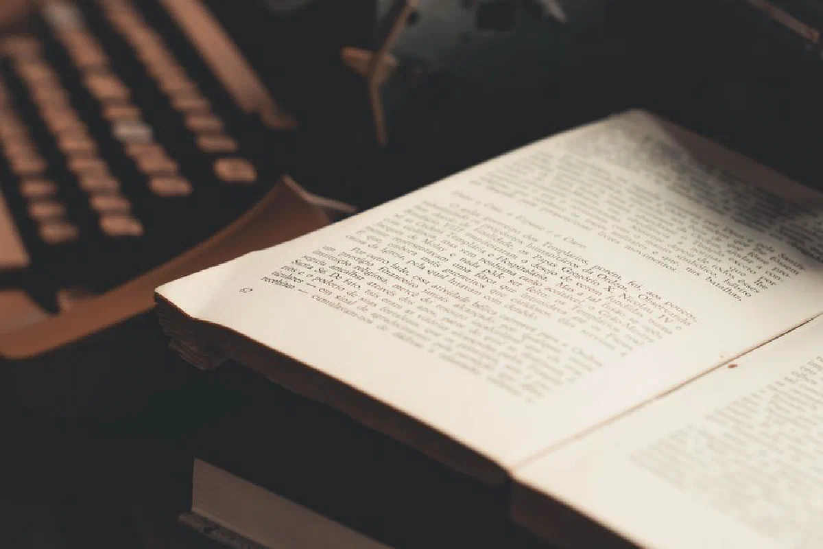 An open book with visible text in front of an antique typewriter, evoking a vintage literary theme.