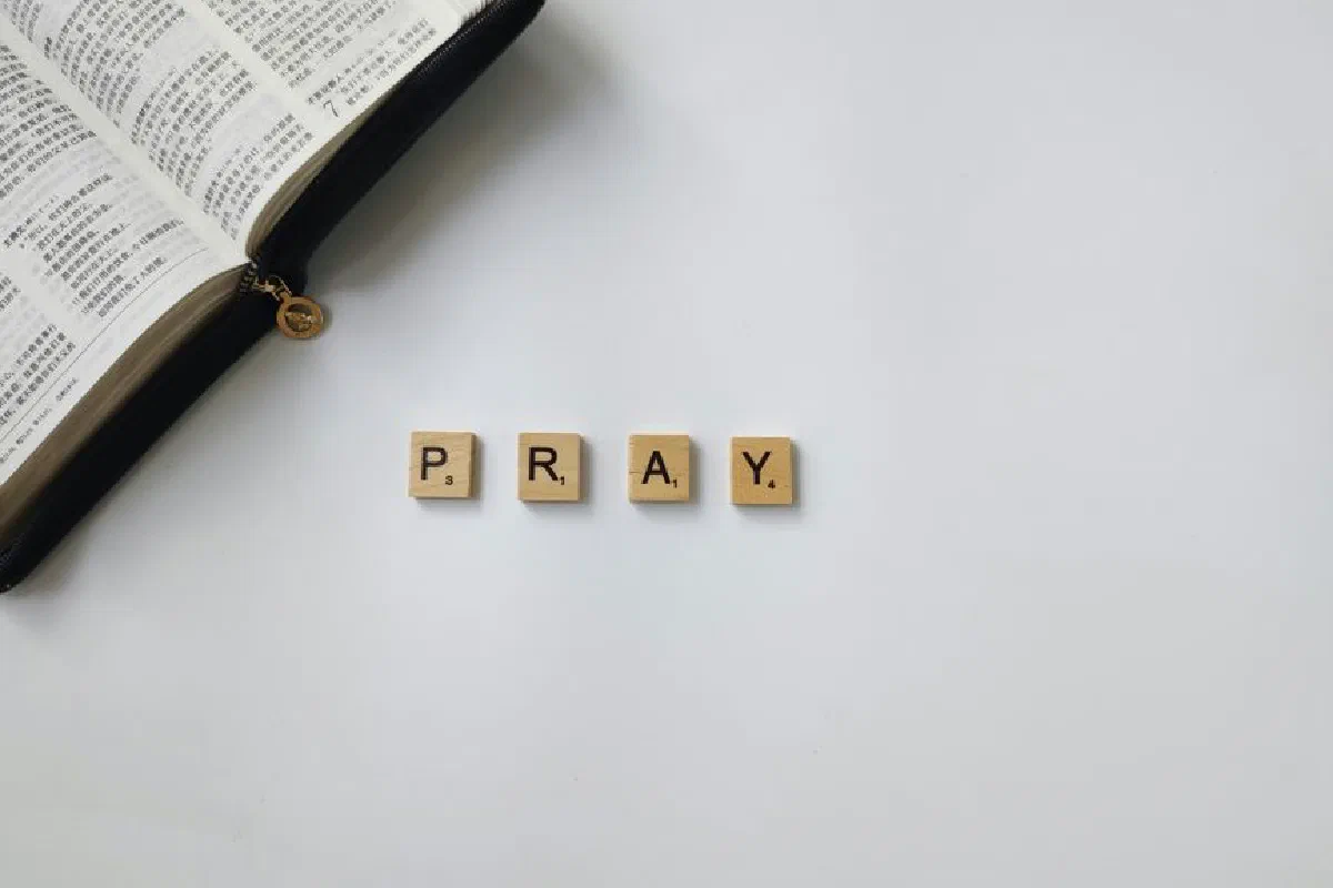 Flat lay of an open Bible and letter tiles spelling 'Pray' on a white background, symbolizing religious reflection.